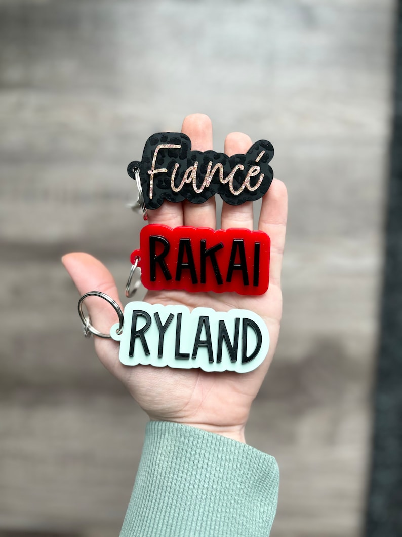 Op de afbeelding: Sleutelhangers met namen en het woord "Fianc&eacute;". De sleutelhangers zijn in zwart, rood en lichtgroen. Het woord "Fianc&eacute;" is in een zwart luipaardprint ontwerp met gouden letters. De andere sleutelhangers hebben de namen "RAKAI" en "RYLAND".