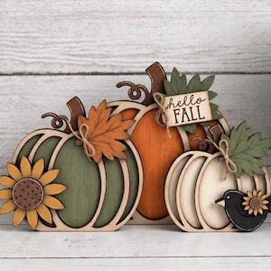 May include: A decorative wooden tabletop display featuring three layered pumpkins in olive green, orange, and cream. The arrangement includes attached wooden sunflowers, maple leaves, a small black bird, and a rectangular sign that reads hello FALL.