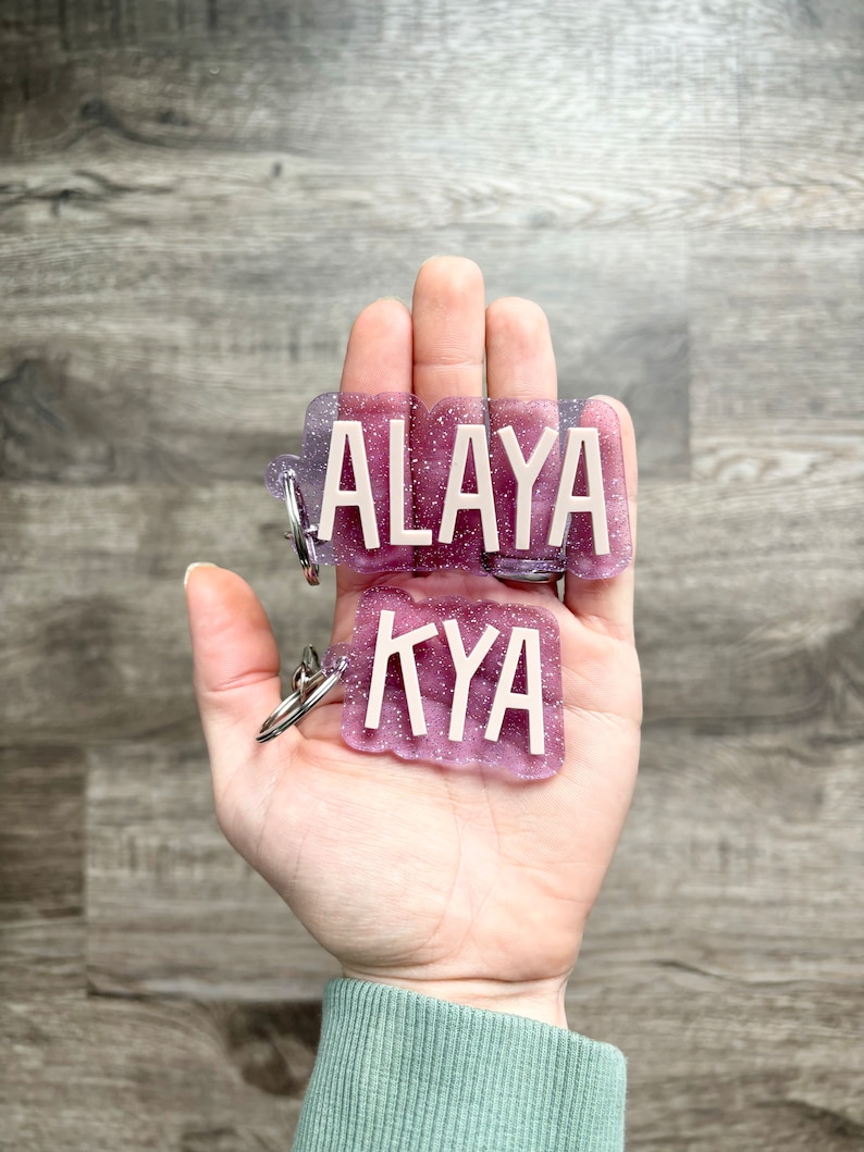 Op de afbeelding: Twee gepersonaliseerde sleutelhangers met de namen "ALAYA" en "KYA" in witte blokletters. De sleutelhangers hebben een glitterende paarse en lavendelkleurige achtergrond. Elke sleutelhanger heeft een zilveren sleutelring.