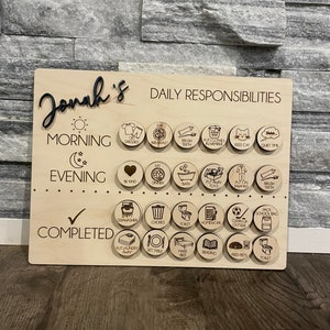 May include: A wooden board with the name "Jonah's" at the top. The board is divided into sections for morning, evening, and completed tasks. Each section has a series of circular icons representing daily responsibilities, such as getting dressed, brushing teeth, eating breakfast, and putting away toys.