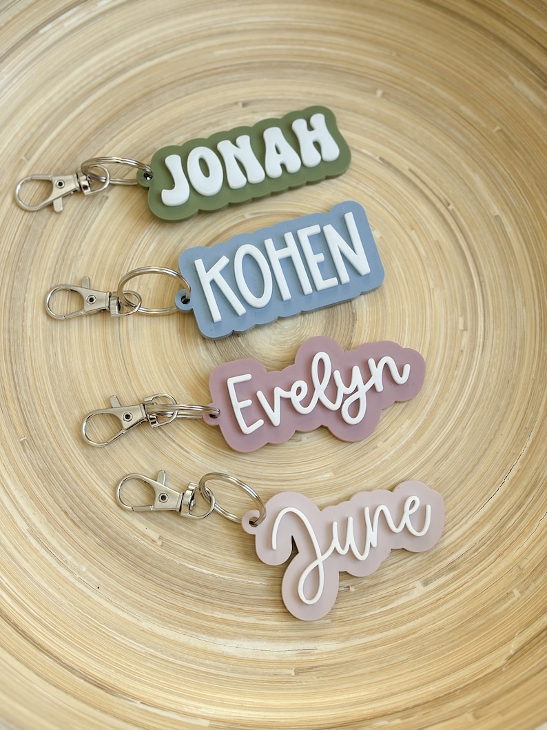 Op de afbeelding: Vier gepersonaliseerde sleutelhangers met namen: "Jonah" in wit op een groene achtergrond, "Kohen" in wit op blauw, "Evelyn" in wit op mauve en "June" in wit op roze. Elke sleutelhanger heeft een zilveren sluiting en ring.