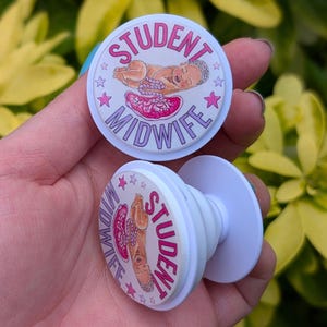 May include: Two white phone grips with a circular design. The design features a baby, a uterus, and the words "STUDENT MIDWIFE" in pink. The grips are held in a hand against a background of green and yellow leaves.
