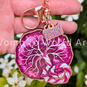 May include: A wooden keychain with a pink and white galaxy design featuring a stylized womb and the text "Womb to World".