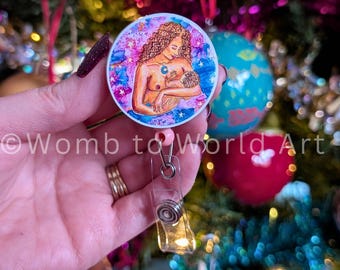 May include: A pink and blue watercolor badge reel with a woman and child depicted in a motherly embrace. The woman has brown skin and long brown hair. The child is nestled in her arms. The background is a watercolor wash of pink, blue, and white with stars.