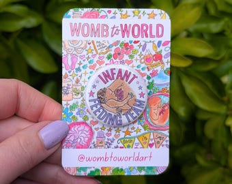 Könnte beinhalten: Ein weißer Knopf mit einer bunten Illustration einer Person, die ein Baby stillt. Der Knopf trägt die Aufschrift "Infant Feeding Team" in einem rosa Kreis. Der Knopf befindet sich auf einer Karte mit einem bunten Hintergrund aus Blumen, Sternen und anderen Illustrationen. Die Karte trägt außerdem die Aufschrift "Womb to World" und "@wombtoworldart".
