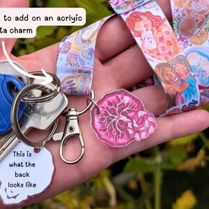 May include: A fabric lanyard featuring a colorful illustration of people and fetuses, attached to a metal keyring with a blue key and a pink acrylic placenta charm. The charm has a white back with the text: This is what the back looks like.