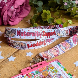 Può includere: Un cordino in tessuto bianco con un motivo ripetuto di illustrazioni raffiguranti neonati e la scritta Maternity Support in lettere rosse. Il cordino include un moschettone girevole in metallo argentato fissato a un porta badge in plastica rosa.