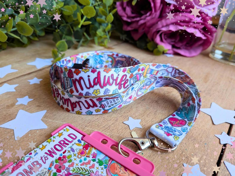 May include: A white fabric lanyard featuring a colorful repeating pattern of medical icons, flowers, and the word midwife in pink script. The lanyard includes a silver metal swivel lobster clasp attached to a bright pink plastic ID badge holder.