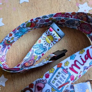 May include: A white fabric lanyard featuring a colorful repeating pattern of medical and baby-themed illustrations, including a sun, clogs, rainbows, and flowers. The word Midwife is printed in pink script text along the strap, which includes a black plastic buckle and a metal attachment ring.
