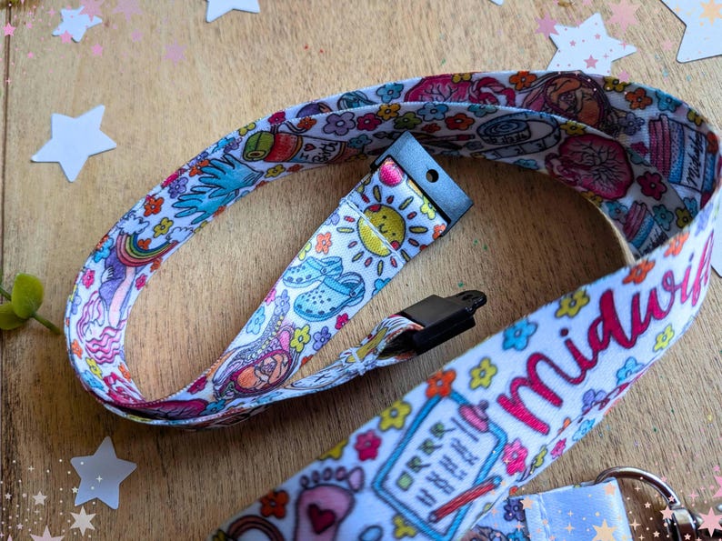 May include: A white fabric lanyard featuring a colorful repeating pattern of medical and baby-themed illustrations, including a sun, clogs, rainbows, and flowers. The word Midwife is printed in pink script text along the strap, which includes a black plastic buckle and a metal attachment ring.