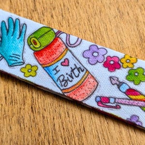 May include: A white fabric strap featuring a colorful pattern of teal medical gloves, a red and orange water bottle with the text I Heart Birth, various small multi-colored flowers, and a rainbow-striped pen with a pink cap and a fountain pen nib.