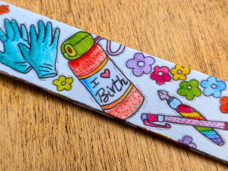 May include: A white fabric strap featuring a colorful pattern of teal medical gloves, a red and orange water bottle with the text I Heart Birth, various small multi-colored flowers, and a rainbow-striped pen with a pink cap and a fountain pen nib.
