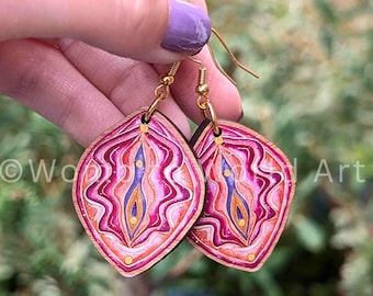 May include: A pair of wooden earrings with a pink, purple, and gold abstract design. The earrings are shaped like a diamond and have gold hooks.