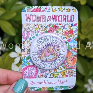 May include: A white button with a purple border and the text "COMMUNITY HEALTH WORKER" in black. The button features a cartoon illustration of a person with purple hair holding a baby. The button is on a card with a pink background and colourful illustrations of flowers, hearts, and other birth-related imagery. The text "WOMB to WORLD" is at the top of the card.