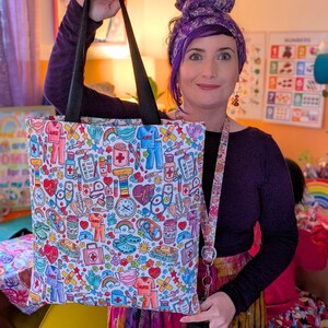 May include: A white tote bag with a colorful print of medical symbols, including hearts, bandages, stethoscopes, and pills. The bag has black handles and a rainbow-colored strap.