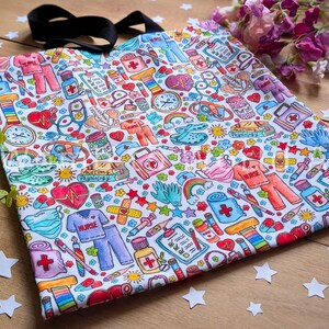 May include: A white tote bag with a colorful pattern of medical-themed illustrations. The pattern includes hearts, bandages, pills, syringes, stethoscopes, and the word "NURSE".