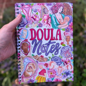 May include: Spiral-bound notebook with a colorful cover featuring illustrations of pregnancy, childbirth, and flowers. The words "DOULA Notes" are written in a decorative font. The notebook is held against a blurred green and purple background.