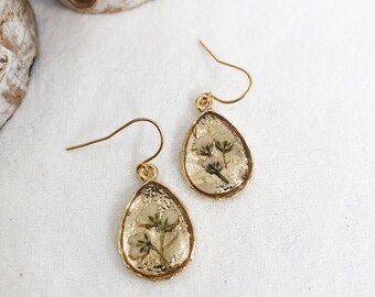 May include: A pair of gold-toned teardrop-shaped earrings featuring pressed white flowers encased in a textured resin setting. Each earring hangs from a simple gold-toned hook wire, showcasing a botanical design with a metallic finish.