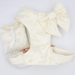 May include: A white dog harness with a large white satin bow and pearl embellishments. The harness has a matching white pillow with pearl embellishments.
