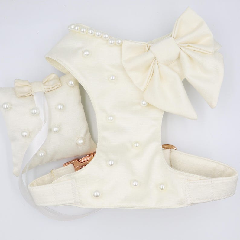 May include: A white dog harness with a large white satin bow and pearl embellishments. The harness has a matching white pillow with pearl embellishments.
