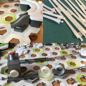 Pu&ograve; includere: Primo piano di un gioco di biglie con una variet&agrave; di piste, rampe e ostacoli. Il gioco &egrave; realizzato in plastica bianca e grigia e presenta un tabellone esagonale verde e bianco. Il gioco include una variet&agrave; di pezzi di gioco di biglie, tra cui una rampa grigia, una rampa bianca, un anello grigio, un anello bianco, una pista grigia, una pista bianca, un interruttore grigio, un interruttore bianco, una biglia grigia, una biglia bianca e una biglia verde.