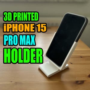 May include: A white 3D printed phone holder designed for an iPhone 15 Pro Max. The holder is angled to prop up the phone for viewing.