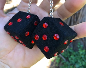 Op de afbeelding: Twee zwarte dobbelstenen met rode edelsteenstippen, verbonden door een zilveren ketting. De dobbelstenen zijn gemaakt van een zacht materiaal. De achtergrond is een vage groene en bruine kleur.