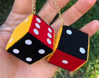 Op de afbeelding: Oorbellen met dobbelstenen. Elke dobbelsteen is een kubus met zwarte, rode en gele zijden en witte stippen. De dobbelstenen hangen aan een goudkleurige ketting. De oorbellen worden in een hand gehouden tegen een vage groene achtergrond.