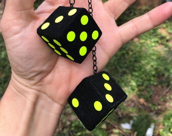 Op de afbeelding: Twee zwarte dobbelstenen met gele stippen die aan een zwarte ketting hangen. De dobbelstenen zijn pluche en hebben een zachte textuur.