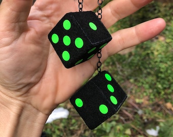 Op de afbeelding: Twee zwarte dobbelstenen met groene stippen die aan een zwarte ketting hangen. De dobbelstenen zijn pluche en hebben een zachte textuur.