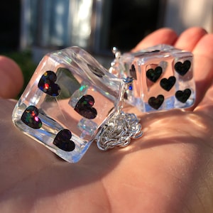 May include: Clear resin dice pendant with black heart-shaped glitter inclusions. The dice are connected by a silver chain. The dice are held in a hand, with sunlight in the background.