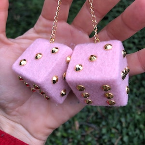 Op de afbeelding: Twee roze dobbelstenen van vilt met gouden steentjes, hangend aan een gouden ketting.