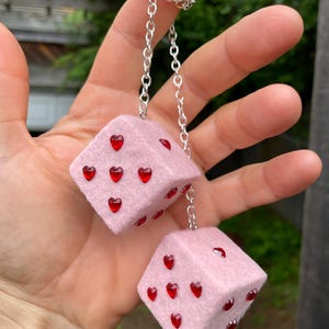 May include: Pair of pink dice with red heart-shaped embellishments. The dice are connected by a silver chain. The dice are held up by a hand.
