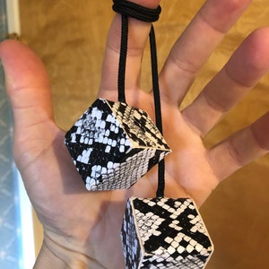 Pu&ograve; includere: Due dadi neri e bianchi con motivo a pelle di serpente sono attaccati a uno spago nero. Lo spago &egrave; legato attorno a un dito.