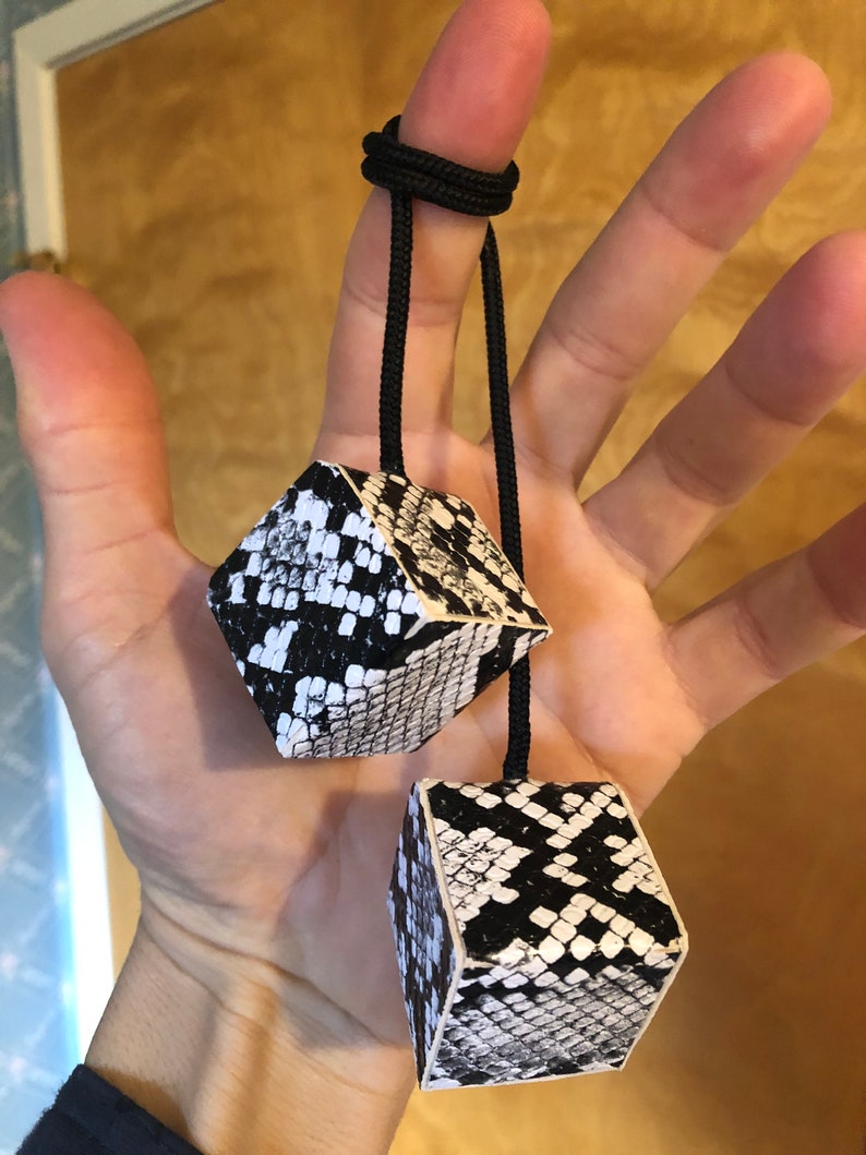 Pu&ograve; includere: Due dadi neri e bianchi con motivo a pelle di serpente sono attaccati a uno spago nero. Lo spago &egrave; legato attorno a un dito.