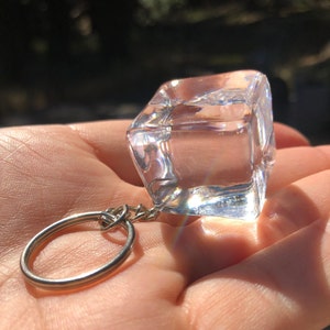 Può includere: Un portachiavi di cristallo trasparente e sfaccettato a forma di cubo. Il portachiavi ha un anello e una catena in metallo color argento. Il cristallo riflette la luce, creando un effetto scintillante. Lo sfondo è sfocato.