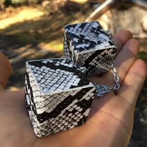 Pu&ograve; includere: Due cubi con motivo a pelle di serpente in bianco e nero collegati da una catena argentata.