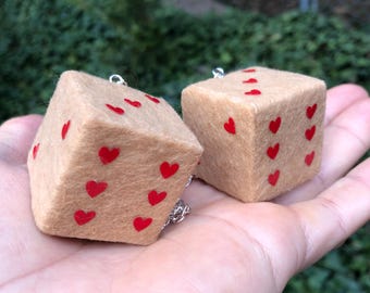 Op de afbeelding: Twee bruinbeige dobbelstenen met rode hartvormige markeringen. De dobbelstenen zijn gemaakt van een zacht materiaal en zijn verbonden door een zilveren ketting. De dobbelstenen worden in de hand van een persoon gehouden tegen een achtergrond van groen gebladerte.
