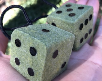 Op de afbeelding: Twee groene vilten dobbelstenen met zwarte stippen, elk bevestigd aan een zwart touwtje. De dobbelstenen bevinden zich in een hand, met de groene vilten kant naar de camera gericht.