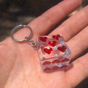 Peut inclure: Un porte-clés en résine transparente en forme de dé avec des cœurs rouges pailletés sur chaque face. Le porte-clés a un anneau en métal argenté.