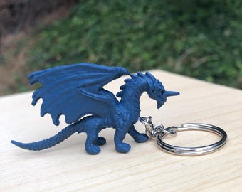 May include: A blue dragon keychain with a silver key ring. The dragon is posed in a standing position with its wings spread out.
