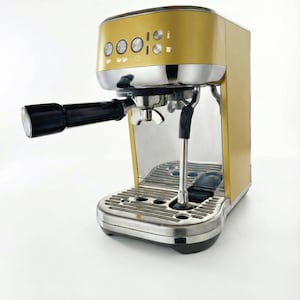 Op de afbeelding: Een goud-zilveren espressomachine met een zwarte handgreep en bedieningsknoppen. De machine heeft een roestvrijstalen lekbak en waterreservoir. Ontworpen voor het maken van espresso en andere koffiedranken. Afmetingen zijn ongeveer 30 cm lang.