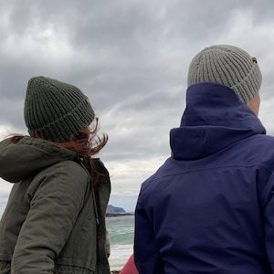 以下が含まれることがあります： 2 人が冬用の帽子とジャケットを着て、曇り空を背景に水面に向かって立っています。