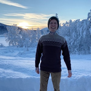 Puede incluir: Un hombre con un suéter de punto marrón y beige con un patrón, pantalones de pana marrón y un gorro verde con un parche marrón. Está de pie en un paisaje nevado con árboles en el fondo.