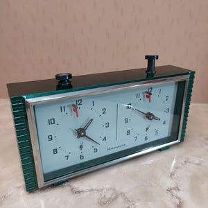 May include: A vintage green and silver chess clock with two clock faces. Each face has black numerals, red hands, and the word "Anmaps" is printed on the face. The clock has two black winding knobs on top.