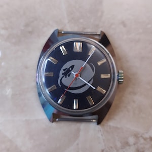 Op de afbeelding: Een vintage polshorloge met een donkerblauwe wijzerplaat met een raketontwerp in een grijze cirkel. Het horloge heeft goudkleurige uurmarkeringen en zilveren wijzers, met een rode secondewijzer. De horlogekast is zilverkleurig.