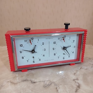May include: A vintage red chess clock with two white clock faces, black hands, and a silver frame. The clock has two black knobs on top and the word "Janfaz" is printed on the face. The clock is marked "MADE IN USSR".