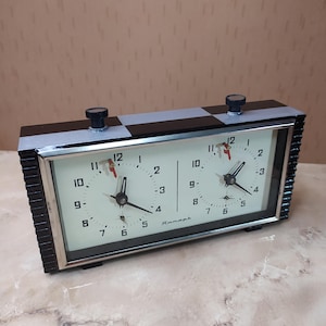 May include: A rectangular analog chess clock with a black and silver casing. The device features two white clock faces with black numerals, black hands, and red indicator flags. The word Yantar is printed in Cyrillic script on the clock face.
