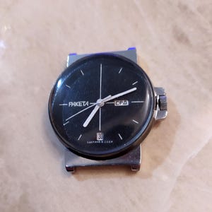 Puede incluir: Un reloj de pulsera Raketa vintage con esfera negra, manecillas plateadas y caja plateada. La esfera muestra el día de la semana y el número 30. Un reloj clásico.