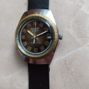 Puede incluir: Un reloj de pulsera de oro y plata con una correa de cuero negro. La esfera del reloj es marrón con detalles dorados y el texto "POLJOT 23 jewels" y "MADE IN USSR". La ventana de la fecha muestra el número 28.